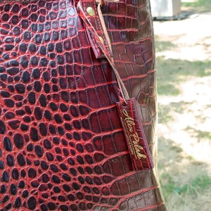 May include: A deep red tote bag with a glossy crocodile skin texture. The bag features two shoulder straps attached with gold-toned metal rivets and a rectangular leather tag hanging from a strap, embossed with the text Alex Polak.