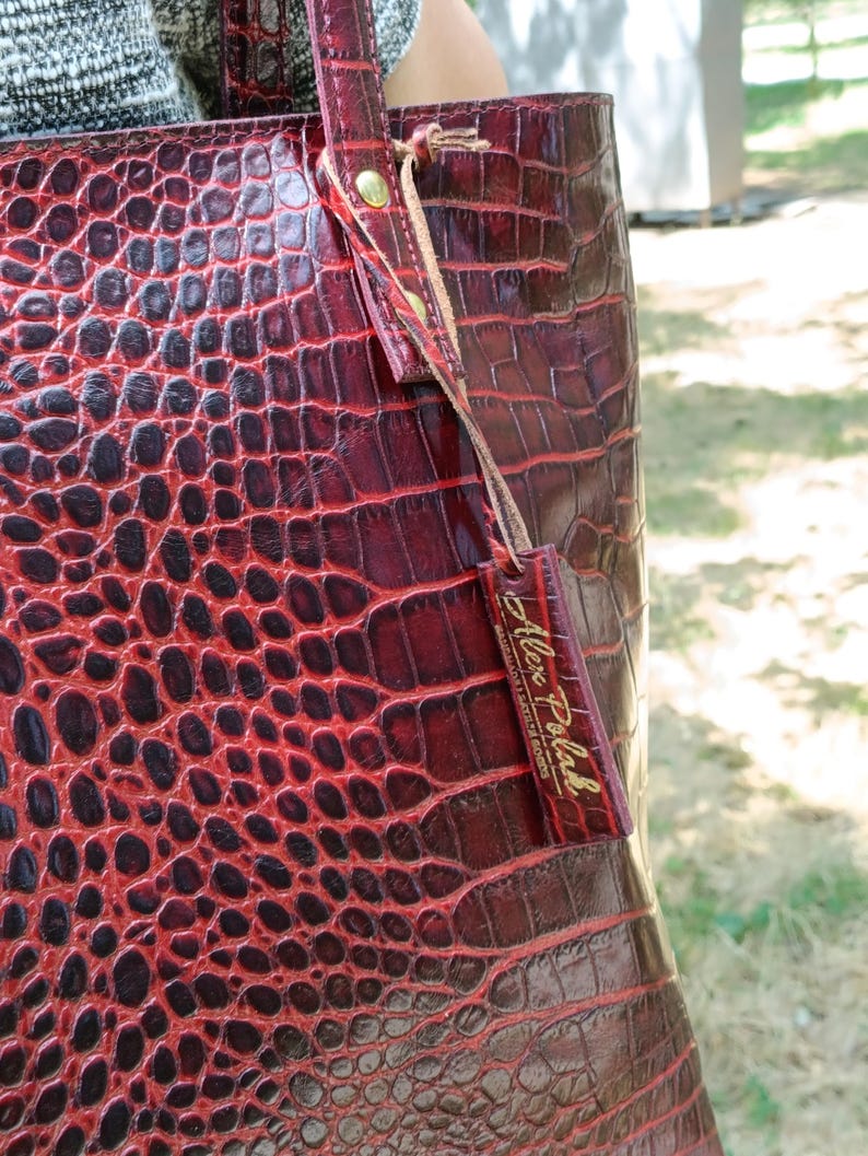 May include: A deep red tote bag with a glossy crocodile skin texture. The bag features two shoulder straps attached with gold-toned metal rivets and a rectangular leather tag hanging from a strap, embossed with the text Alex Polak.