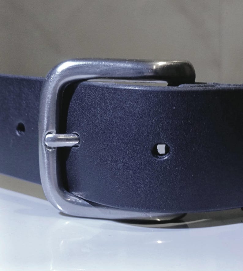 Op de afbeelding: Close-up van een marineblauwe leren riem met een zilverkleurige gesp. De riem heeft een rechthoekige gesp en meerdere gaten voor verstelling. Het leer ziet er glad uit en de gesp glanst.