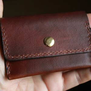 May include: A small, brown leather wallet with a brass snap closure. The wallet is rectangular with rounded corners and features visible stitching in a lighter brown thread. The leather has a rich, textured appearance.