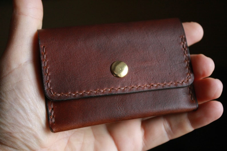 May include: A small, brown leather wallet with a brass snap closure. The wallet is rectangular with rounded corners and features visible stitching in a lighter brown thread. The leather has a rich, textured appearance.