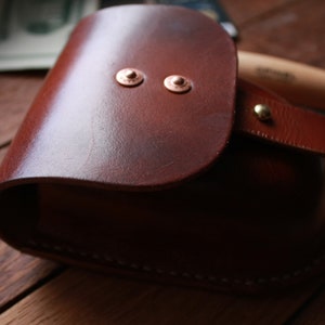 May include: A brown leather pouch with a flap closure and two copper rivets. The pouch has a white stitching detail around the edges.