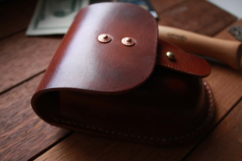 May include: A brown leather pouch with a flap closure and two copper rivets. The pouch has a white stitching detail around the edges.