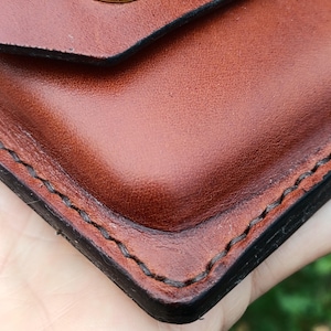 Puede incluir: Un primer plano de una cartera de cuero marr&oacute;n con costuras oscuras visibles a lo largo de los bordes y un cierre de bot&oacute;n a presi&oacute;n de metal dorado pulido. El cuero tiene una textura suave con bordes oscuros bru&ntilde;idos y una esquina redondeada.