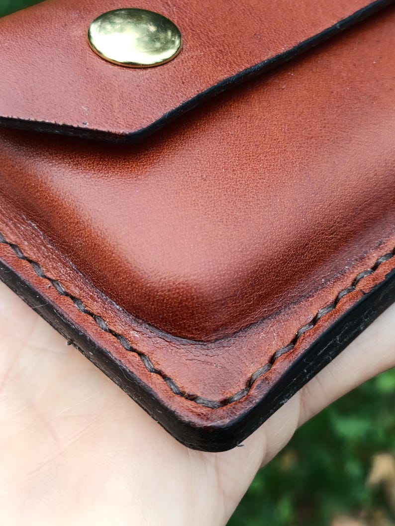 Puede incluir: Un primer plano de una cartera de cuero marr&oacute;n con costuras oscuras visibles a lo largo de los bordes y un cierre de bot&oacute;n a presi&oacute;n de metal dorado pulido. El cuero tiene una textura suave con bordes oscuros bru&ntilde;idos y una esquina redondeada.