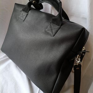 Puede incluir: Un malet&iacute;n de cuero negro con una superficie texturizada. El bolso tiene un asa superior y una correa para el hombro desmontable. La cremallera recorre la parte superior y lateral del bolso. El bolso est&aacute; sobre una superficie blanca.