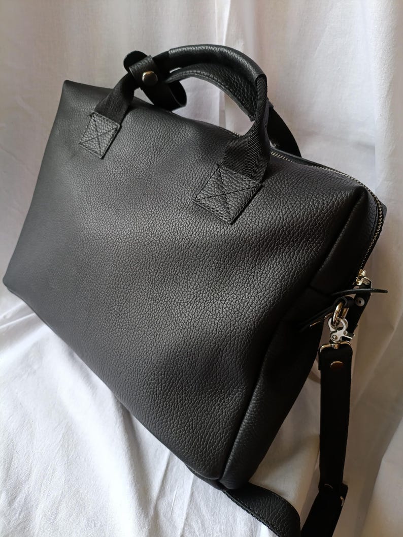 Puede incluir: Un malet&iacute;n de cuero negro con una superficie texturizada. El bolso tiene un asa superior y una correa para el hombro desmontable. La cremallera recorre la parte superior y lateral del bolso. El bolso est&aacute; sobre una superficie blanca.