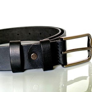 K&ouml;nnte beinhalten: Nahaufnahme eines schwarzen Lederg&uuml;rtels mit einer silberfarbenen rechteckigen Schnalle. Der G&uuml;rtel hat ein klassisches Design mit einer einzelnen Schlaufe und einem Metallknopfverschluss. Der G&uuml;rtel befindet sich auf einer reflektierenden Oberfl&auml;che.