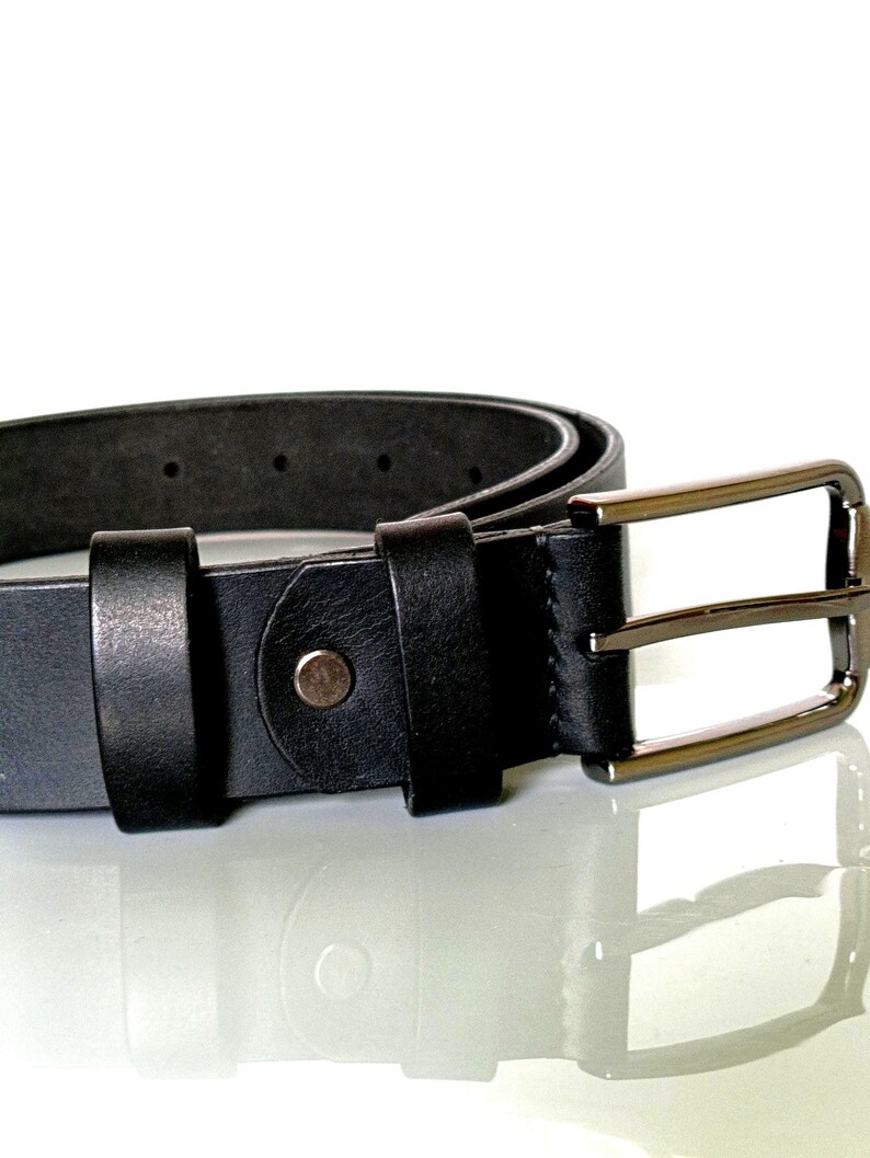 K&ouml;nnte beinhalten: Nahaufnahme eines schwarzen Lederg&uuml;rtels mit einer silberfarbenen rechteckigen Schnalle. Der G&uuml;rtel hat ein klassisches Design mit einer einzelnen Schlaufe und einem Metallknopfverschluss. Der G&uuml;rtel befindet sich auf einer reflektierenden Oberfl&auml;che.
