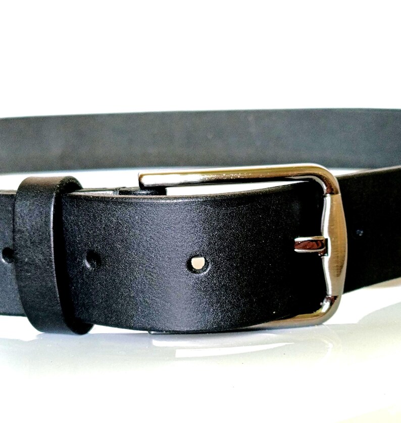 K&ouml;nnte beinhalten: Nahaufnahme eines schwarzen Lederg&uuml;rtels mit silberner Schnalle. Der G&uuml;rtel hat ein klassisches Design mit rechteckiger Schnalle und einer einzelnen G&uuml;rtelschlaufe. Das Leder wirkt glatt und ist f&uuml;r den t&auml;glichen Gebrauch geeignet.