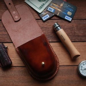 May include: A brown leather pouch with a flap and a snap closure. The pouch is sitting on a wooden surface with a pocket knife, a lighter, and a pocket watch.