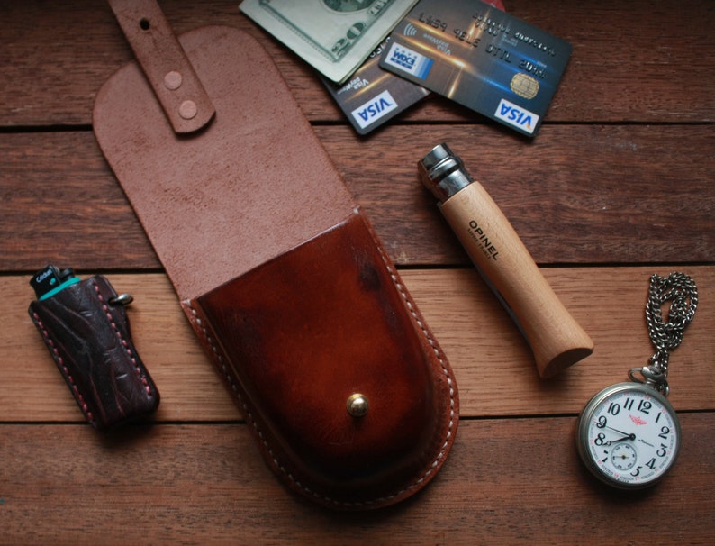 May include: A brown leather pouch with a flap and a snap closure. The pouch is sitting on a wooden surface with a pocket knife, a lighter, and a pocket watch.
