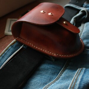 May include: A brown leather pouch with a flap closure and metal snaps. The pouch is attached to a belt and is sitting on a pair of blue jeans.
