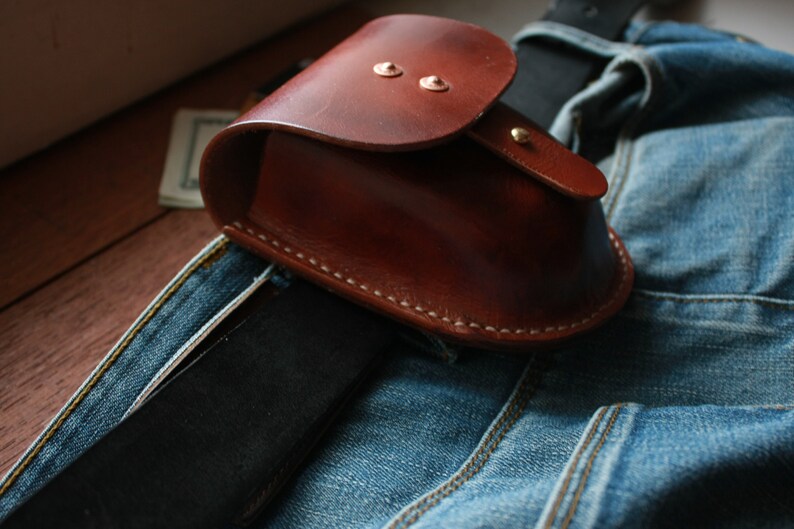 May include: A brown leather pouch with a flap closure and metal snaps. The pouch is attached to a belt and is sitting on a pair of blue jeans.