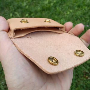 May include: A light tan leather pouch featuring a fold-over flap with two brass snap fasteners. The interior compartment is open, revealing the natural texture of the leather. Two matching brass studs are visible on the top edge of the pouch.
