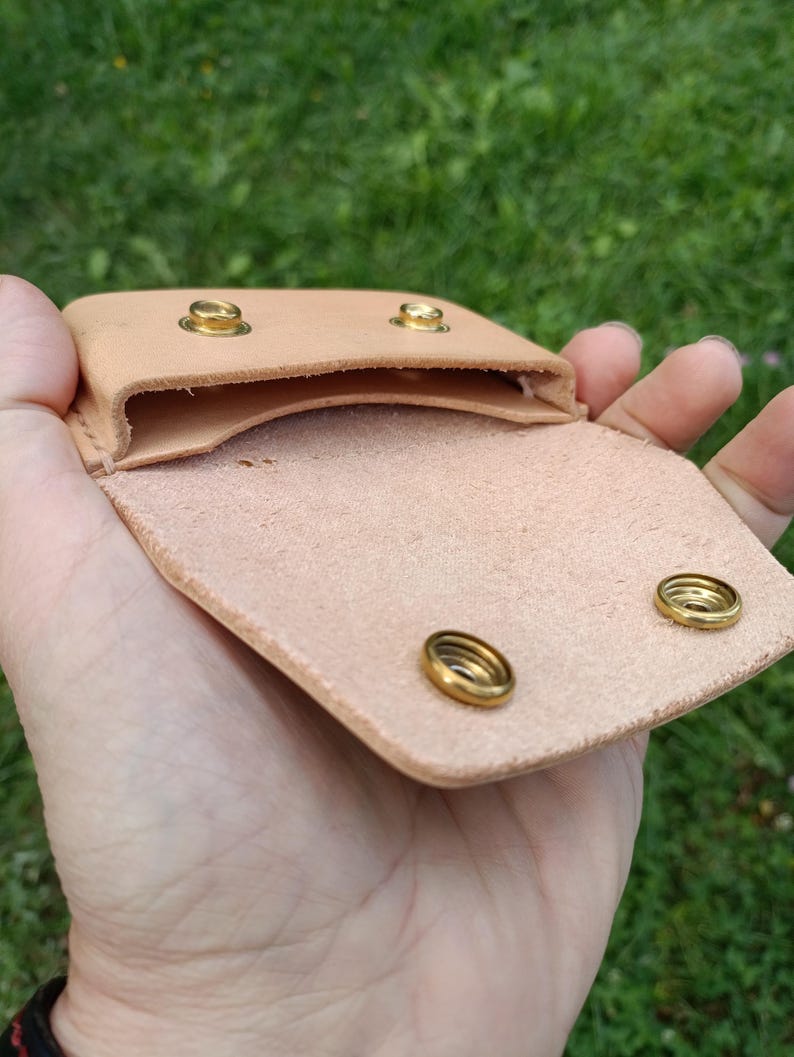 May include: A light tan leather pouch featuring a fold-over flap with two brass snap fasteners. The interior compartment is open, revealing the natural texture of the leather. Two matching brass studs are visible on the top edge of the pouch.