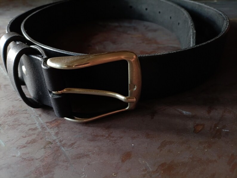 Puede incluir: Un cintur&oacute;n de cuero negro con un acabado mate liso y una hebilla de metal en tono dorado pulido. El cintur&oacute;n incluye una trabilla de cuero a juego y una serie de agujeros preperforados a lo largo de la tira para un ajuste seguro.