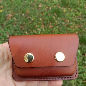 May include: A small, rectangular brown leather pouch featuring a fold-over flap secured by two polished gold-tone metal snap buttons. The edges of the pouch are finished with visible dark brown stitching along the perimeter.