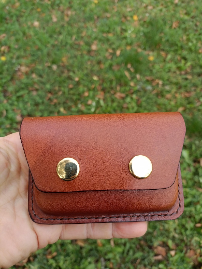 May include: A small, rectangular brown leather pouch featuring a fold-over flap secured by two polished gold-tone metal snap buttons. The edges of the pouch are finished with visible dark brown stitching along the perimeter.