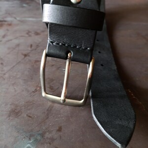 Puede incluir: Un cintur&oacute;n de cuero negro con una hebilla de metal plateado y una trabilla de cuero a juego. La correa del cintur&oacute;n est&aacute; asegurada con un remache de metal plateado cerca de la hebilla e incluye una serie de agujeros perforados a lo largo.