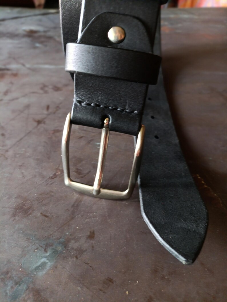 Puede incluir: Un cintur&oacute;n de cuero negro con una hebilla de metal plateado y una trabilla de cuero a juego. La correa del cintur&oacute;n est&aacute; asegurada con un remache de metal plateado cerca de la hebilla e incluye una serie de agujeros perforados a lo largo.