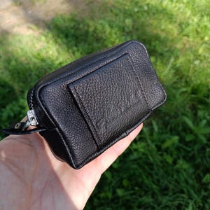 Puede incluir: Un peque&ntilde;o estuche rectangular negro con textura granulada y una cremallera de metal plateado. La parte frontal presenta un bolsillo rectangular con el texto en relieve Alex Bee. El estuche tiene una forma estructurada con costuras visibles.