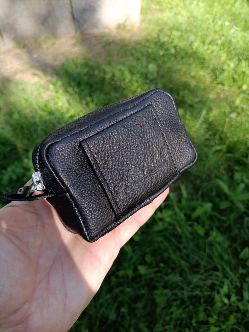 Puede incluir: Un peque&ntilde;o estuche rectangular negro con textura granulada y una cremallera de metal plateado. La parte frontal presenta un bolsillo rectangular con el texto en relieve Alex Bee. El estuche tiene una forma estructurada con costuras visibles.