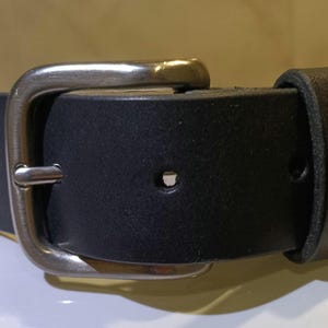 Op de afbeelding: Close-up van een zwarte leren riem met een zilverkleurige gesp. De riem heeft een rechthoekige gesp met een enkele pen. Het leer lijkt glad, geschikt voor dagelijks gebruik. De riem is ongeveer 3-4 cm breed.