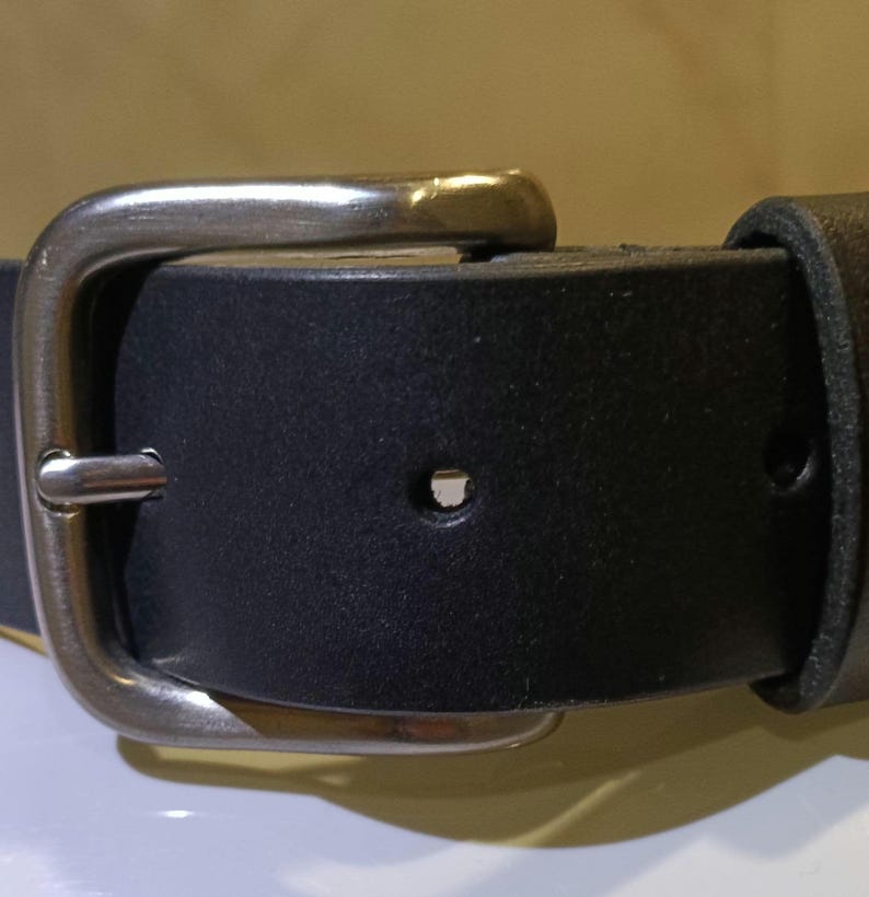 Op de afbeelding: Close-up van een zwarte leren riem met een zilverkleurige gesp. De riem heeft een rechthoekige gesp met een enkele pen. Het leer lijkt glad, geschikt voor dagelijks gebruik. De riem is ongeveer 3-4 cm breed.