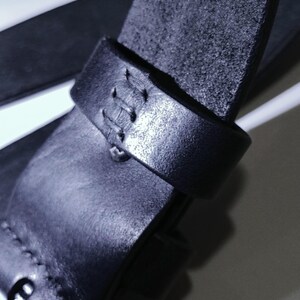 May include: Close-up of a black leather belt with a textured surface. The belt features a stitched loop and a buckle. The leather appears to be of high quality, with visible grain and a matte finish. The belt is designed for everyday wear.