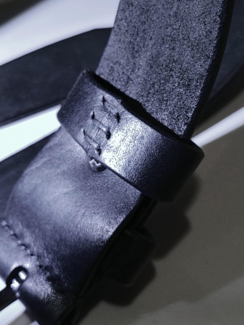May include: Close-up of a black leather belt with a textured surface. The belt features a stitched loop and a buckle. The leather appears to be of high quality, with visible grain and a matte finish. The belt is designed for everyday wear.