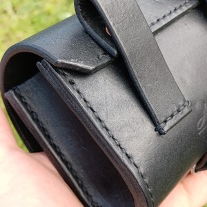 May include: Close-up of a black leather case with a curved design and a strap closure. The case has visible stitching and a smooth, matte finish. The background is blurred green.