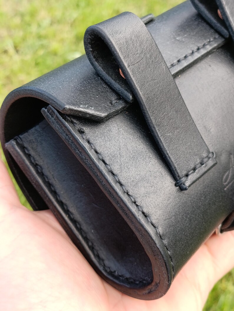 May include: Close-up of a black leather case with a curved design and a strap closure. The case has visible stitching and a smooth, matte finish. The background is blurred green.