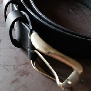 Puede incluir: Un cintur&oacute;n de cuero negro con un acabado mate liso y una hebilla de metal plateado pulido. El cintur&oacute;n incluye dos trabillas de cuero negro a juego cerca de la hebilla, la cual est&aacute; asegurada a la tira con un remache met&aacute;lico visible.