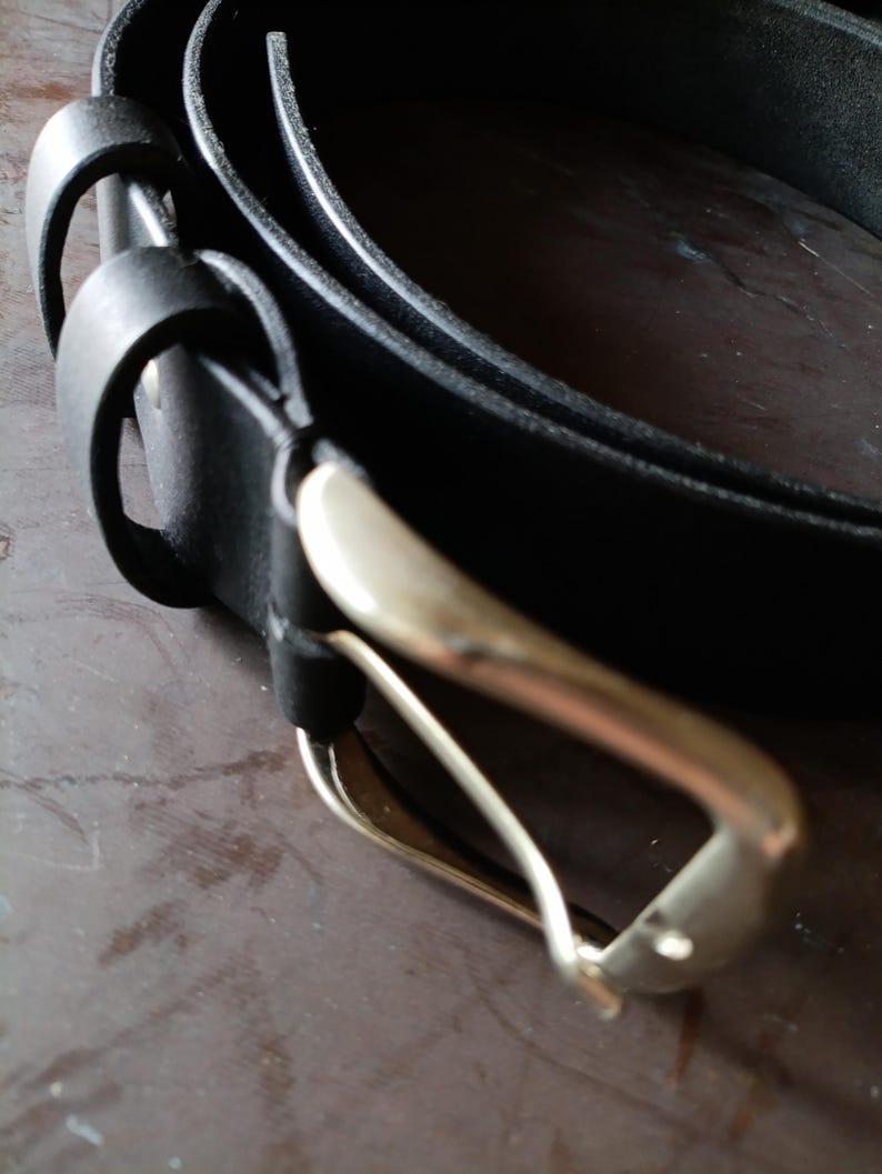 Puede incluir: Un cintur&oacute;n de cuero negro con un acabado mate liso y una hebilla de metal plateado pulido. El cintur&oacute;n incluye dos trabillas de cuero negro a juego cerca de la hebilla, la cual est&aacute; asegurada a la tira con un remache met&aacute;lico visible.