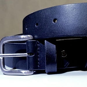 Op de afbeelding: Close-up van een zwarte leren riem met een zilverkleurige gesp. De riem heeft verschillende verstelgaten en een getextureerd oppervlak. De gesp is rechthoekig en heeft een klassiek ontwerp.