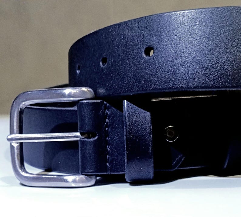 Op de afbeelding: Close-up van een zwarte leren riem met een zilverkleurige gesp. De riem heeft verschillende verstelgaten en een getextureerd oppervlak. De gesp is rechthoekig en heeft een klassiek ontwerp.