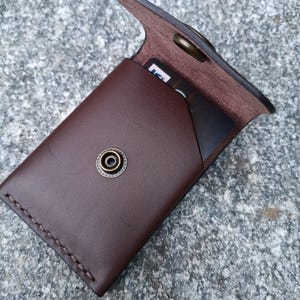 May include: A dark brown leather card holder featuring a flap closure with a brass-toned metal snap button. The wallet has visible stitching along the side and contains a black card with partial white text visible inside the main compartment.