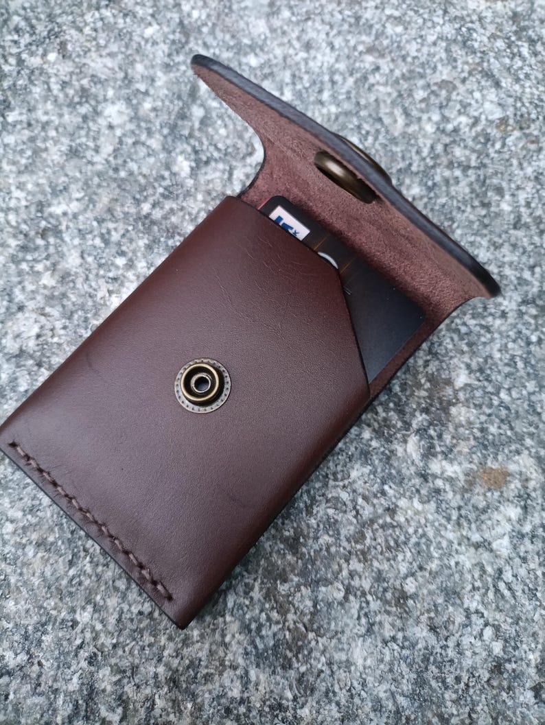 May include: A dark brown leather card holder featuring a flap closure with a brass-toned metal snap button. The wallet has visible stitching along the side and contains a black card with partial white text visible inside the main compartment.