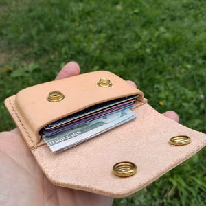 May include: A light tan leather wallet featuring a flap closure secured by two brass snap buttons. The interior compartment holds several stacked credit cards and a folded United States dollar bill, with visible stitching along the edges.