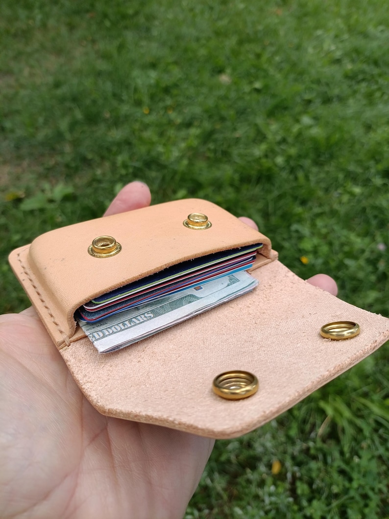 May include: A light tan leather wallet featuring a flap closure secured by two brass snap buttons. The interior compartment holds several stacked credit cards and a folded United States dollar bill, with visible stitching along the edges.