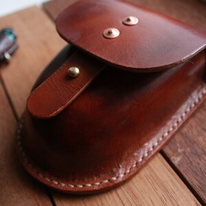 May include: A brown leather pouch with a flap closure and brass accents. The pouch is stitched with white thread and has a curved shape.