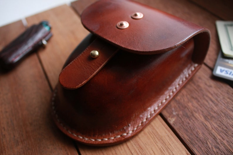May include: A brown leather pouch with a flap closure and brass accents. The pouch is stitched with white thread and has a curved shape.