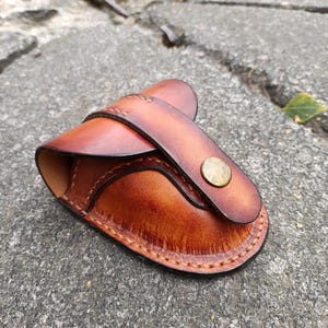 K&ouml;nnte beinhalten: Eine handgefertigte Ledertasche in Braunt&ouml;nen, mit einem geschwungenen Design und einem Riemenverschluss, der mit einem Messingknopf gesichert ist. Die Kanten sind mit einem Kontrastfaden sorgf&auml;ltig gen&auml;ht.