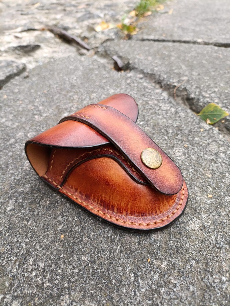 K&ouml;nnte beinhalten: Eine handgefertigte Ledertasche in Braunt&ouml;nen, mit einem geschwungenen Design und einem Riemenverschluss, der mit einem Messingknopf gesichert ist. Die Kanten sind mit einem Kontrastfaden sorgf&auml;ltig gen&auml;ht.