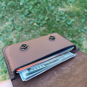 Puede incluir: Una cartera de cuero marr&oacute;n con cierre de solapa asegurado por dos botones de presi&oacute;n de metal oscuro. El compartimento interior contiene varias tarjetas de cr&eacute;dito apiladas y un billete de cien d&oacute;lares estadounidense doblado.