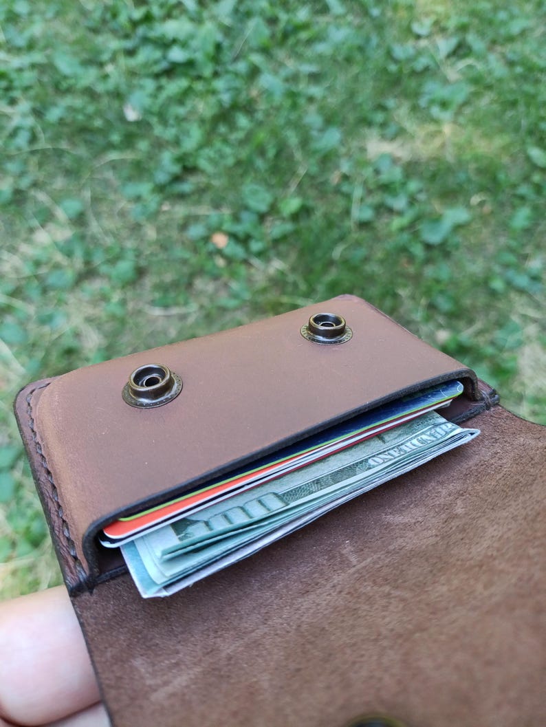 Puede incluir: Una cartera de cuero marr&oacute;n con cierre de solapa asegurado por dos botones de presi&oacute;n de metal oscuro. El compartimento interior contiene varias tarjetas de cr&eacute;dito apiladas y un billete de cien d&oacute;lares estadounidense doblado.