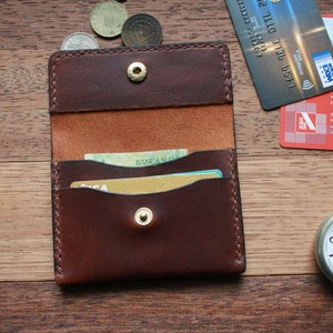 May include: A brown leather card holder with a snap closure, open to reveal cards inside. A Visa credit card and a red credit card are next to the card holder. A silver pocket watch and coins are also on the wooden surface.