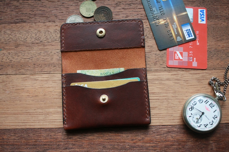 May include: A brown leather card holder with a snap closure, open to reveal cards inside. A Visa credit card and a red credit card are next to the card holder. A silver pocket watch and coins are also on the wooden surface.