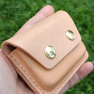 May include: A light tan leather pouch featuring a fold-over flap secured by two polished gold-tone metal snap buttons. The rectangular accessory is constructed with visible stitching along the edges and has a smooth, natural finish throughout.